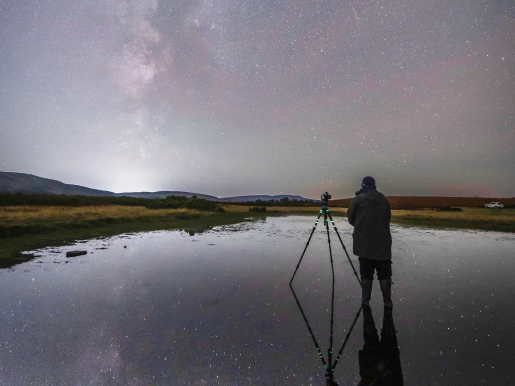 Photographer and tripod beneath the Milky Way reflected in still water