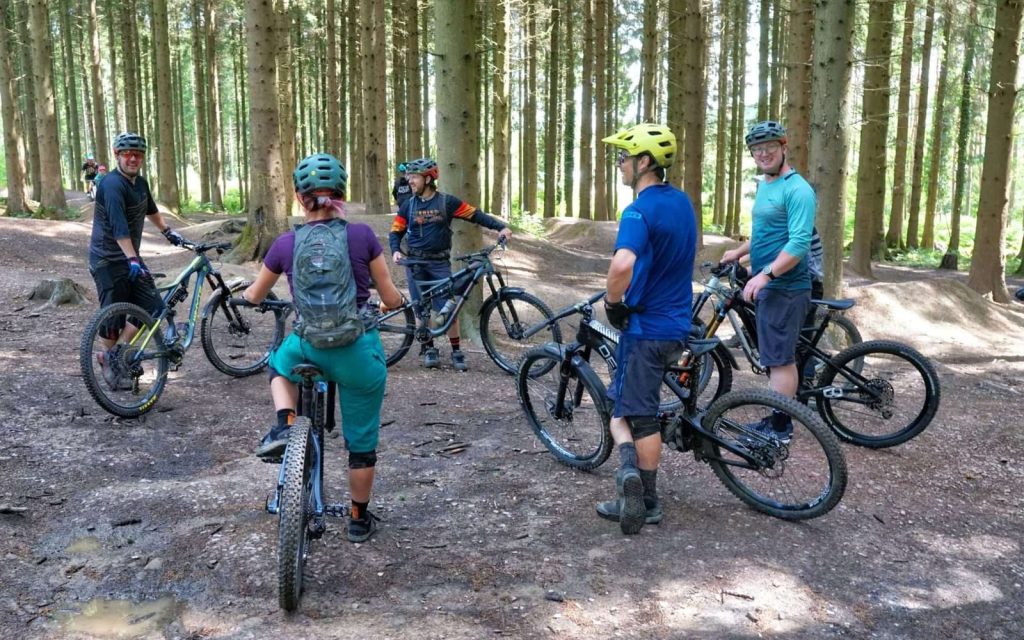 Group of mountain bikers and a coach paused among tall trees with Wye MTB